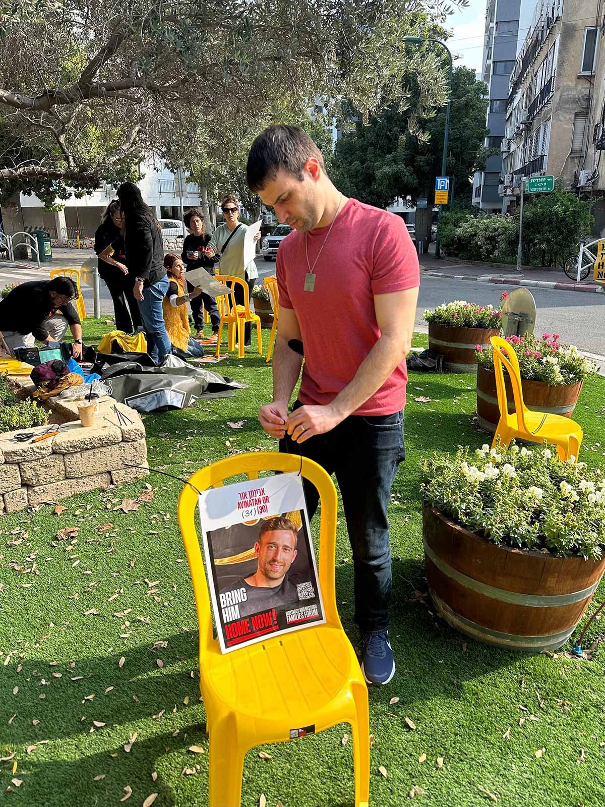 כיכר למען החטופים בשכונת חרוזים ברמת גן. בחור קושר תמונה של חטוף לכסא פלסטיק צהוב. ברקע מספר תושבים מסדרים את כסאות הפלסטיק הצהובים בכיכר.