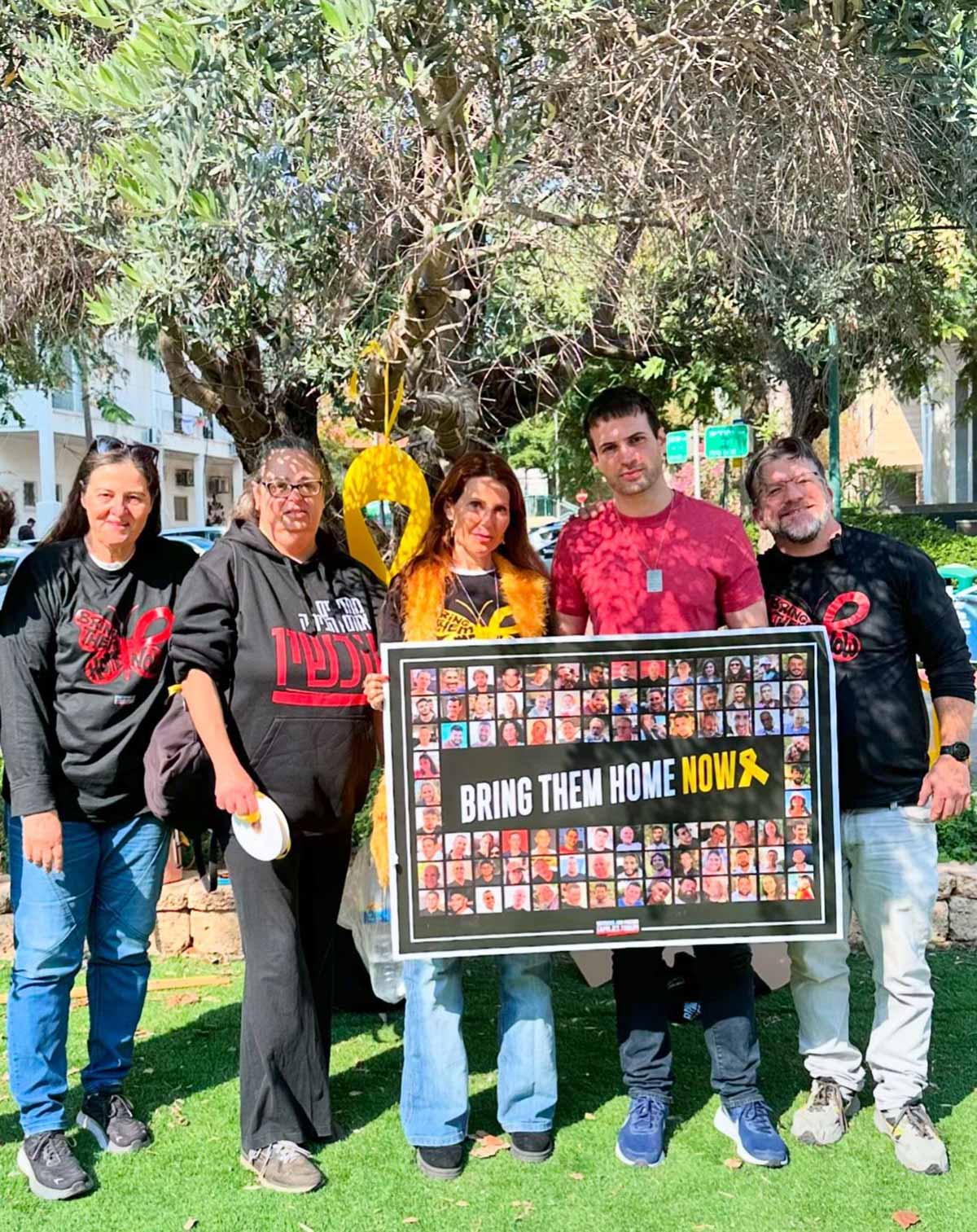 כיכר למען החטופים בשכונת חרוזים ברמת גן. חמישה נשים וגברים עומדים בכיכר עם שלט גדול ובו תמונות החטופים. במרכז השלט כיתוב באנגלית: BRING THEM HOME NOW. מאחוריהם עץ ענק השתול בכיכר.