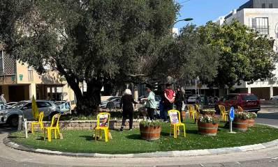כיכר למען החטופים בשכונת חרוזים ברמת גן. תושבים מסדרים כסאות פלסטיק צהובים עם תמונות חטופים בכיכר שיש בה עציצים ועץ ענק. מסביב לכיכר מכוניות ובתי מגורים.