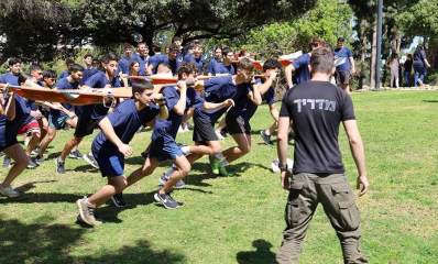 מרוץ הנצחה לבוגרים שנפלו במלחמת חרבות ברזל. נערים ברביעיות אוחזים באלונקות מתחרים בריצה על משטח דשא. לפניהם בצד עומד מדריך.