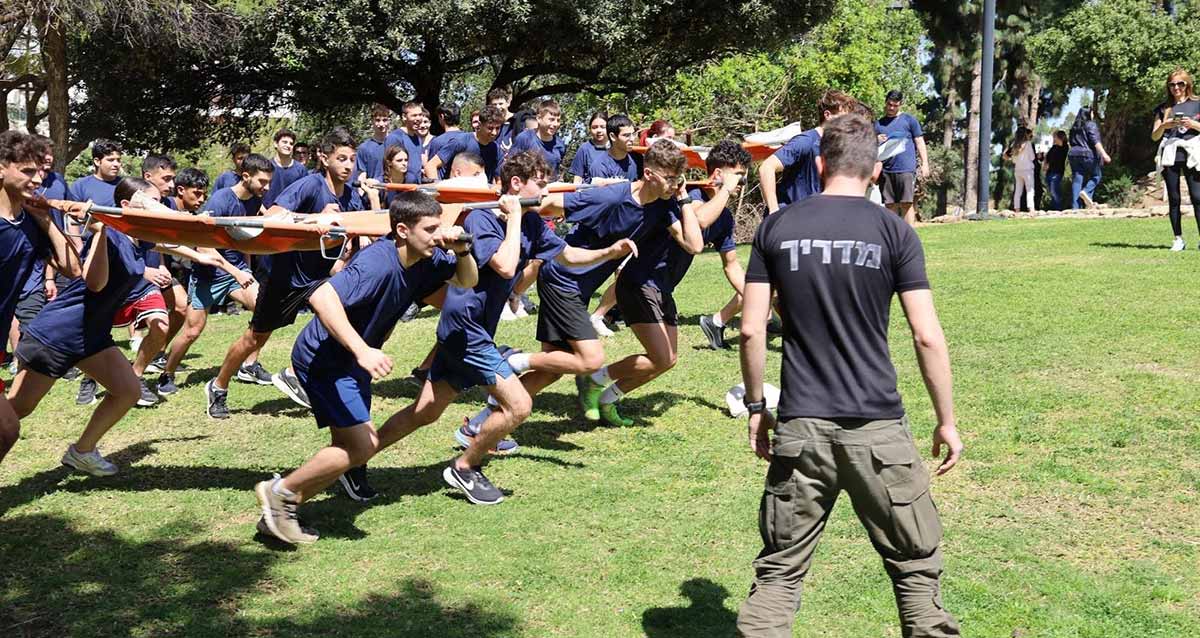 מרוץ הנצחה לבוגרים שנפלו במלחמת חרבות ברזל. נערים ברביעיות אוחזים באלונקות מתחרים בריצה על משטח דשא. לפניהם בצד עומד מדריך.