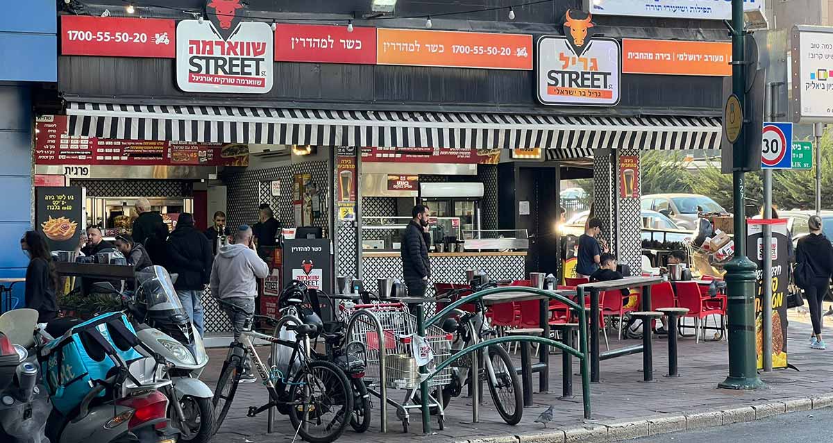 גריל ושווארמה ברחוב ביאליק ברמת גן. קונים בחזית החנות. על המדרכה אופניים ואופנועים. מעל החנות הכיתוב: מעורב ירושלמי, בירה מהחבית, גריל, כשר למהדרין, שווארמה, ומספר טלפון של החנות.