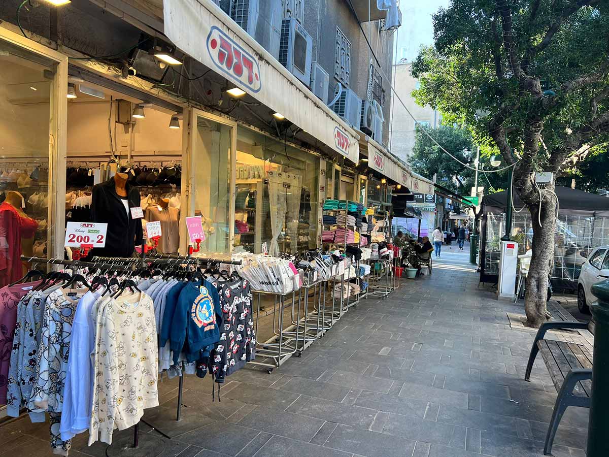 שורה של חנויות ברחוב ביאליק ברמת גן. חנות בגדים בשם דנה. חולצות תלויות על מתלים במדרכה. בגדים על עגלות במדרכה.