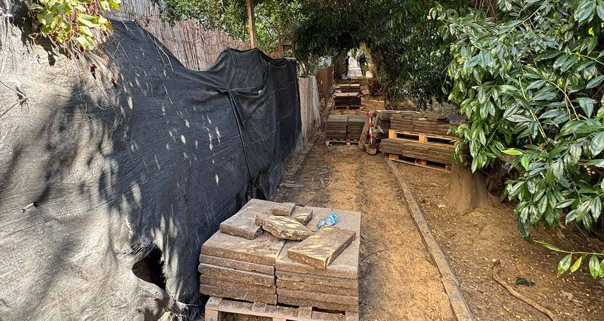 עבודות תשתית בשדרות אפיקים. אבני המדרכה פורקו והן מונחות בערימה. משמאל יריעה שחורה על הגדר של החצר. מימין למדרכה שיחים ועצים.