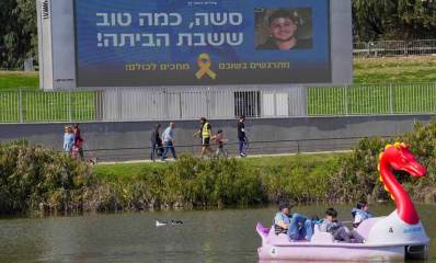 רמת גן חוגגת את חזרתו של סשה טרופנוב. אגם עם סירה בצורת ברבור וילדים ומבוגר יושבים בה. ברקע שביל עם חולכי רגל ומעליהם שלט רחוב גדול עם תמונה של סשה והכיתוב: סשה, כמה טוב ששבת הביתה!