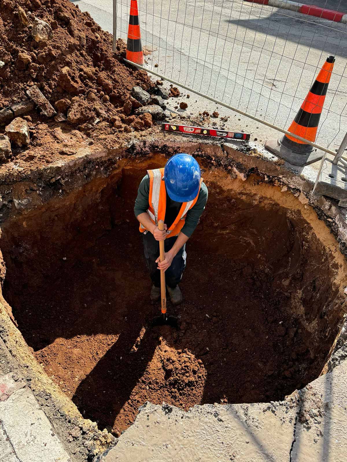 שדרוג תשתית ביוב בויצמן הל"ה. בור עגול ועמוק בכביש בנתיב החסום לתנועה. בתוך הבור פועל חבוש בקסדה עובד עם את חפירה.