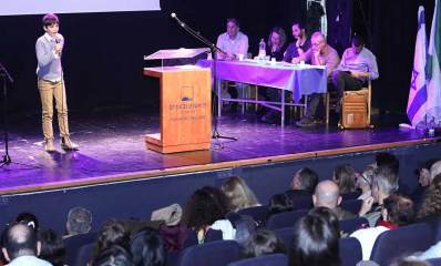 תחרות הנואם הצעיר בגבעתיים. בחור צעיר על הבמה עם מיקרופון ביד. מימין על הבמה שולחן השופטים עם חמישה אנשים. במרכז הבמה דוכן נואמים. מול הבמה ראשים של הקהל.