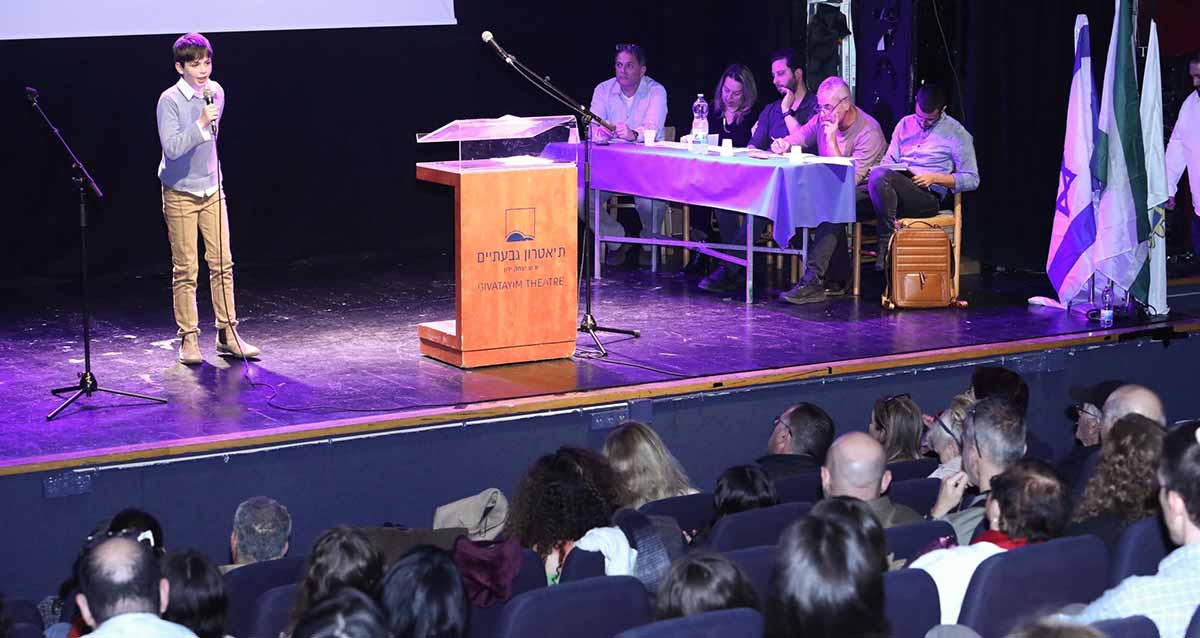 תחרות הנואם הצעיר בגבעתיים. בחור צעיר על הבמה עם מיקרופון ביד. מימין על הבמה שולחן השופטים עם חמישה אנשים. במרכז הבמה דוכן נואמים. מול הבמה ראשים של הקהל.