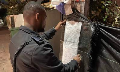 צו סגירה לאתר בנייה בשדרות בן צבי רמת גן. שוטר תולה את שני דפי הצו על גדר מבד שחור, ליד הכניסה לאתר.