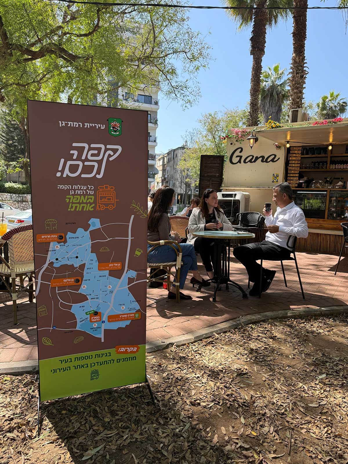 מפה של הפודטראקים ברמת גן ליד פודטראק בשם Gana בגינה ברמת גן. שתי נשים וראש העיר יושבים ליד שולחן עגול ברחבה ליד הפודטראק.