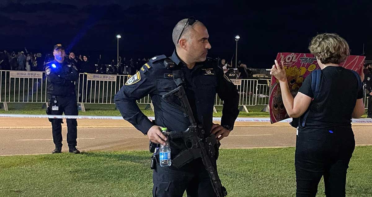 רב פקד לירון לוי, סגן מפקד תחנת תל אביב צפון, עומד ליד מפגינה האוחזת שלט. מאחוריו קצין משטרה. ברקע מפגינים מאחורי שורה של מחסומים על דשא.