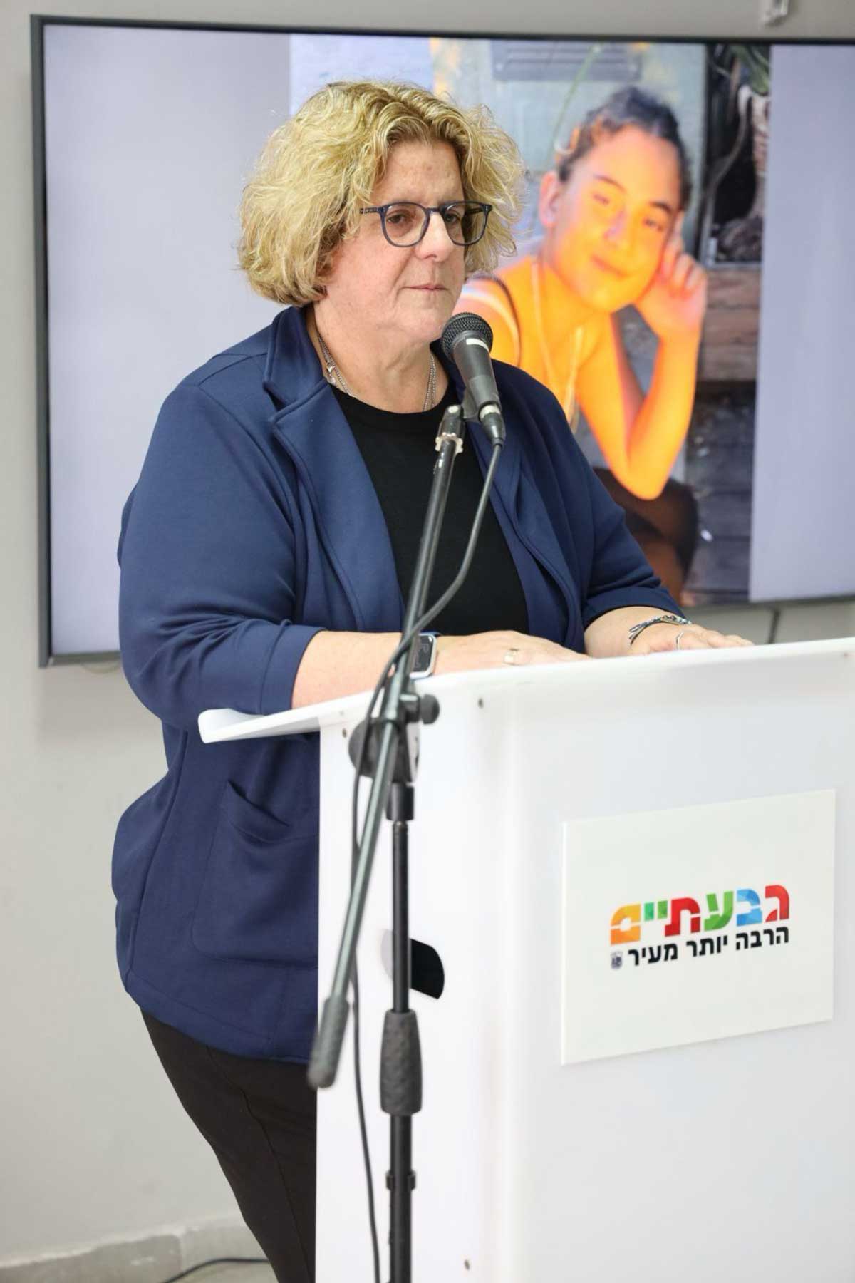 אור-לי ניב, סגנית ראש העיר גבעתיים בתערוכה לזכר התצפיתניות של נחל עוז, ליד דוכן נואמים עם מיקרופון. מאחוריה מוקרנת תמונה של בחורה.