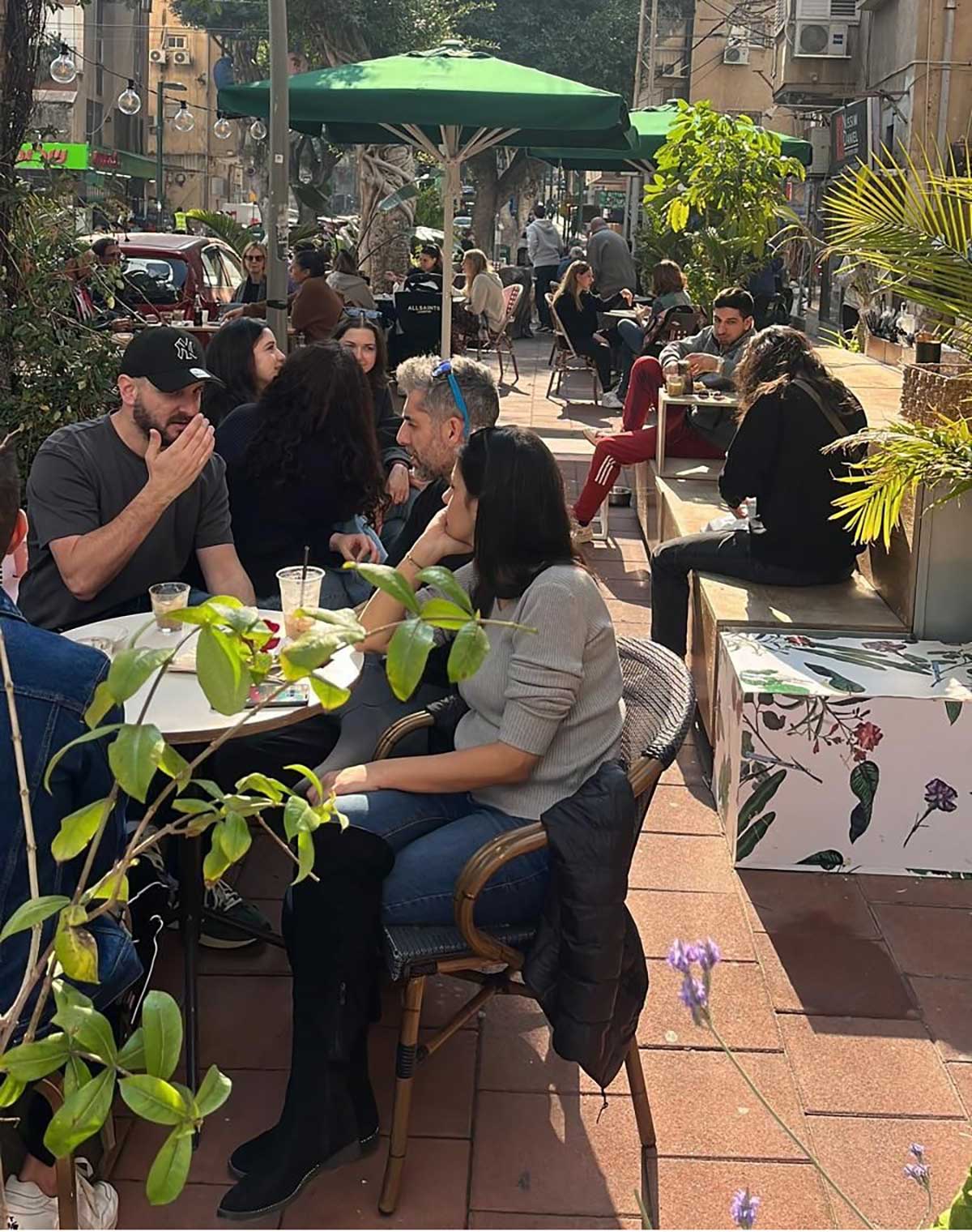 גרימת נזק לבית הקפה "בנק קפה" ברחוב רש"י 23 ברמת גן. אנשים יושבים ברחבה לפני בית הקפה ושמשיות ירוקות מעל.