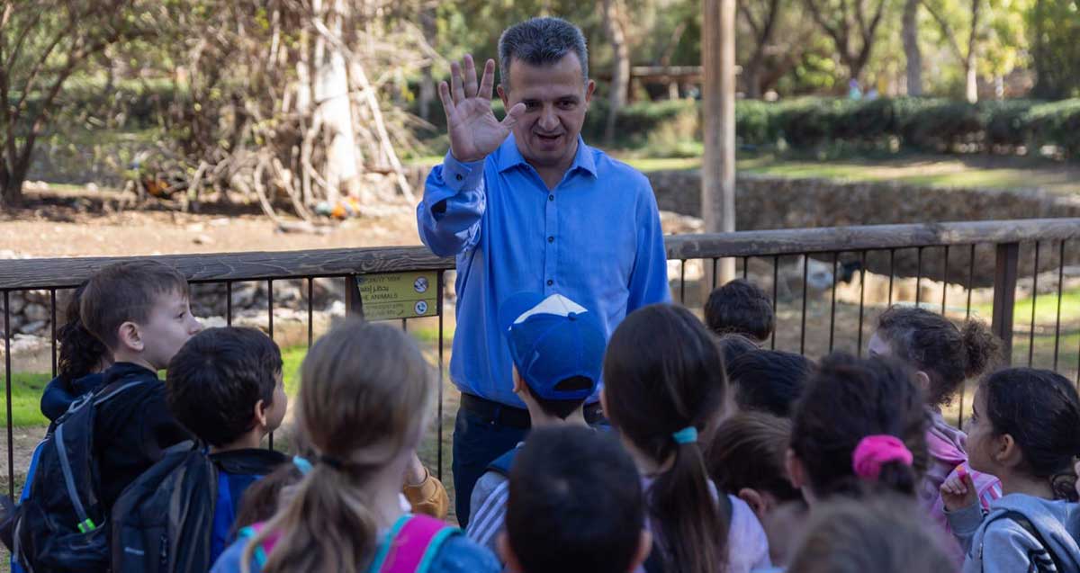 כרמל שאמה הכהן עומד לפני קבוצה של תלמידים ומניף יד לשלום.