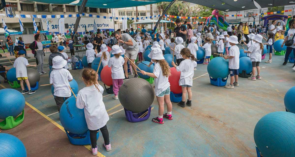 אולימפיאדת גני הילדים ברמת גן. הילדים עומדים בשורות ברחבה עם מקלות ולפניהם כדורים גדולים המונחים על דרגשים.