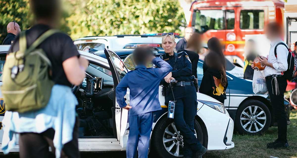 משטרת בני ברק רמת גן באירוע לילדים המתמודדים עם אפילפסיה. ילד בתלבושת כחולה מצדיע לשוטרת הנשענת על מכונית משטרה. רכבים נוספים מאחורי השוטרת. מימין ומשמאל אנשים נוספים עם פנים מטושטשות.