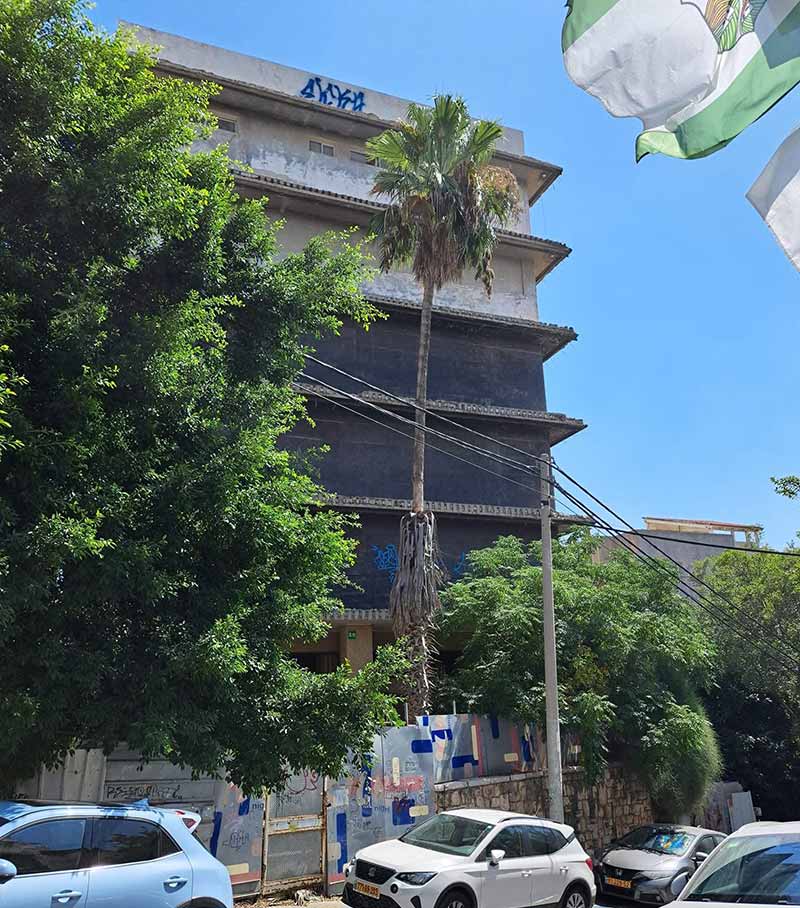 בניין נטוש בגבעתיים. בחזיתו הרחוב ומכוניות חונות. לפניו עצים. בראש הבבניין כתובת גרפיטי כחולה