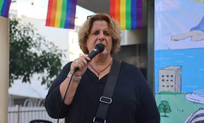אור-לי ניב בחודש הגאווה בגבעתיים עם מיקרופון בידה לפני הפה. מעליה דגלי הגאווה הצבעוניים. מאחוריה כרזה.