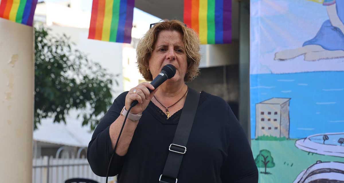 אור-לי ניב בחודש הגאווה בגבעתיים עם מיקרופון בידה לפני הפה. מעליה דגלי הגאווה הצבעוניים. מאחוריה כרזה.