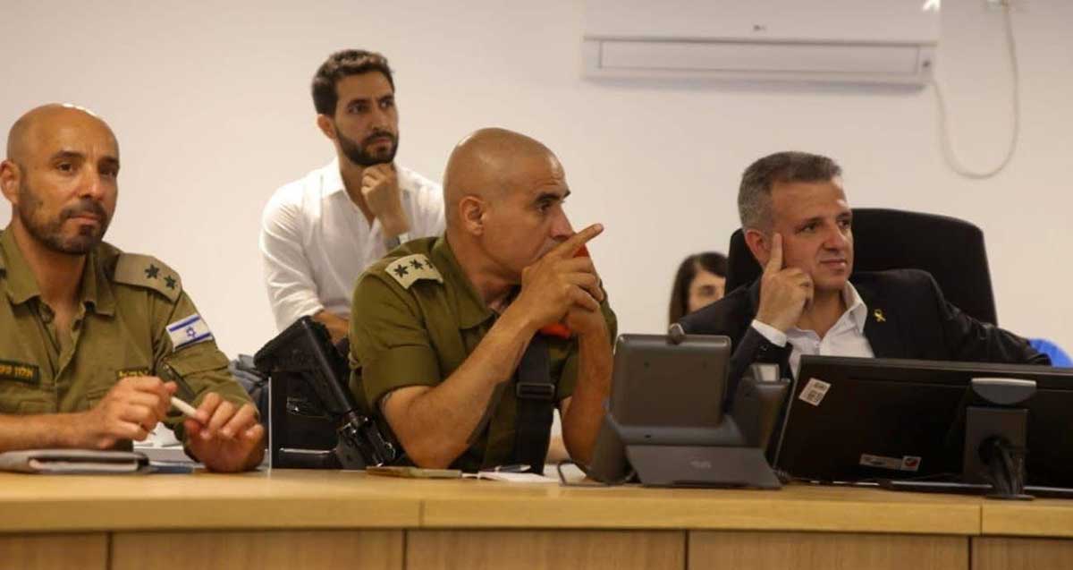 ראש העיר רמת גן במרכז השליטה עם שני קצינים של צה"ל, יושבים ליד שולחן עם מסך, ואיש אחד יושב מאחור.