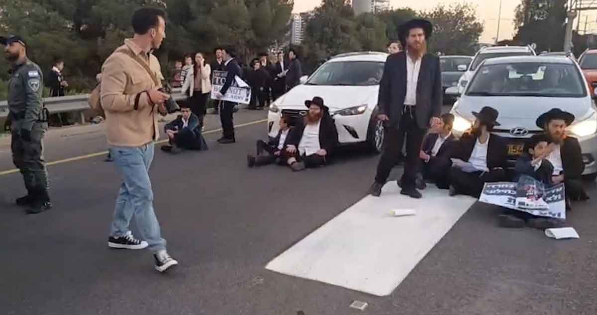 הפגנות וחסימות כבישים בבני ברק. חרדים יושבים על הכביש לפני מכוניות. שוטר עומד בצד.