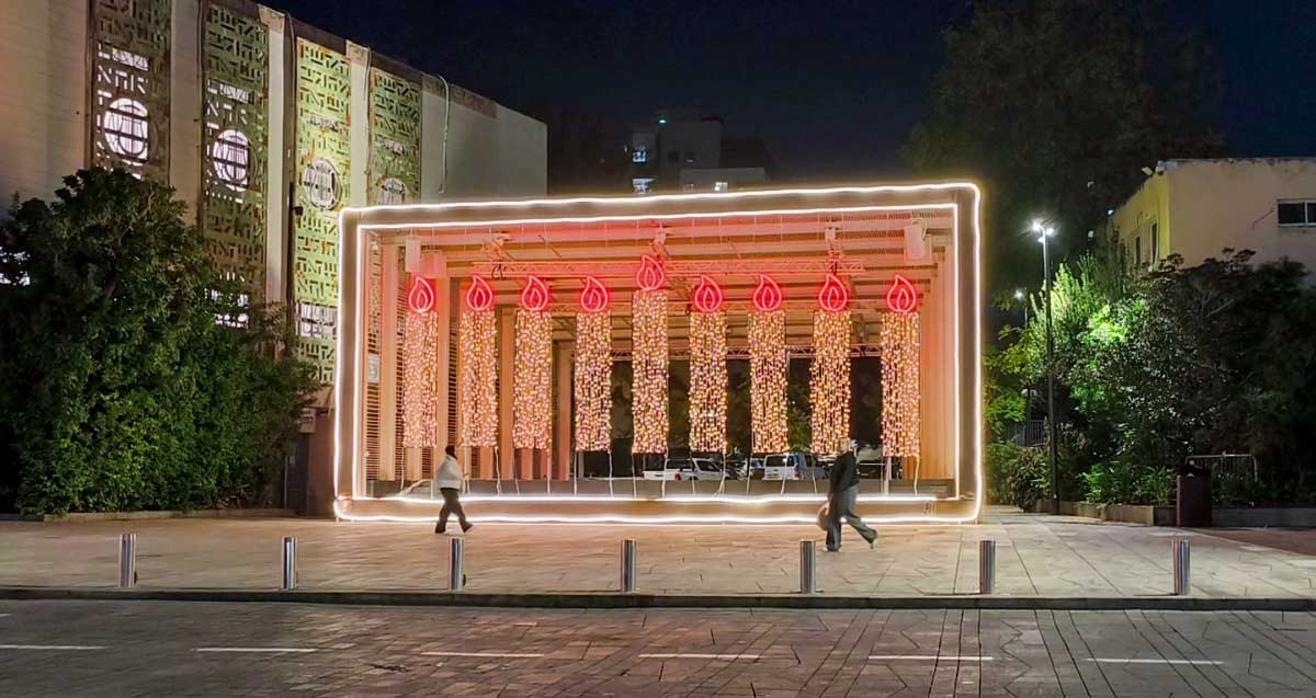 מיצג מואר של חנוכייה ברמת גן ליד בית הכנסת. אנשים צועדים על המדרכה