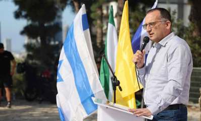 רן קוניק ראש העיר גבעתיים עם מיקרופון ביד עומד מאחורי דוכן נואמים. מאחוריו דגלי המדינה, דגל צהוב ודגל העיר.