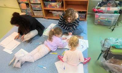 יוזמה לחיבור תלמידי תיכון לילדי הגנים דרך אומנות. שתי תלמידות תיכון עם שתי ילדות על שטיח בגן, מציירות על דפים ברצפה.