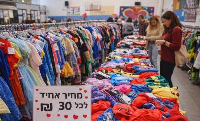 יריד תחפושות לתרומה לחולי סרטן ברמת גן. איור AI. תחפושות על שולחן ארוך ועל מתקן תלייה ארוך. מספר אנשים ליד התחפושות. בחזית התמונה שלט: מחיר אחיד לכל, 30 ש"ח וציורי לבבות אדומים בשלט.