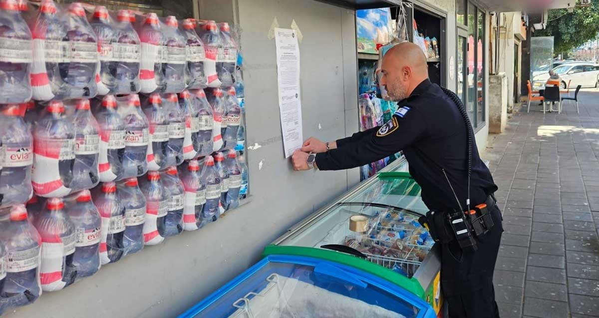 צו סגירה מנהלי למכולת ברמת גן. שוטר מדביק את הצו על הקיר החיצוני של החנות.
