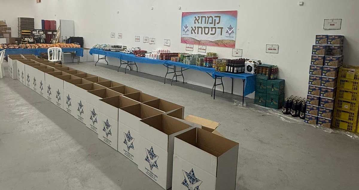 עמותת נחמה והצלה לישראל. אולם עם שולחנות ליד הקירות עם מצרכי מזון לפסח. במרכז האולם שתי שורות של קרטונים פתוחים. על הקיר שלט: רמחא דפסחא.