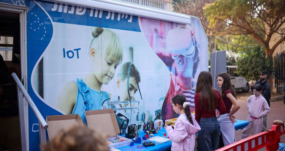 ניידת למידה ברמת גן. ניידת חדשנות עם ציור של ילד וילדה. ליד הניידת שולחן עם מכשירים וילדים ואמהות ליד השולחן.