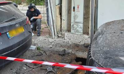 פגיעת רסיס ברמת גן 6.4. בור בין שתי מכוניות מוקף בסרט אדום/לבן וחבלן במדרכה מתבונן בזירה.