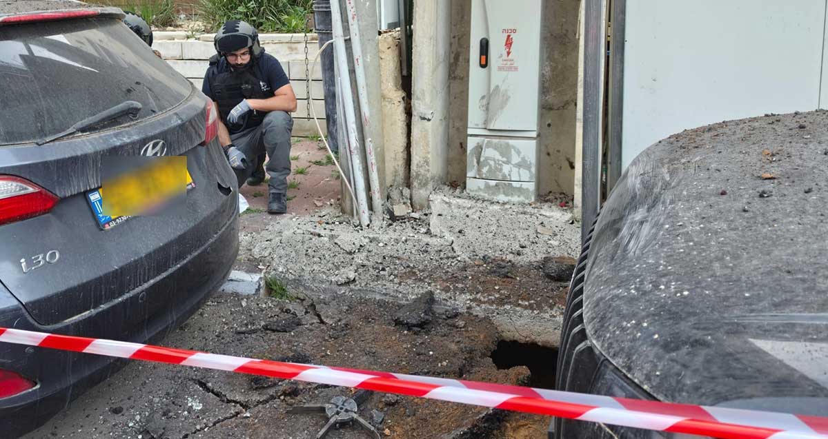 פגיעת רסיס ברמת גן 6.4. בור בין שתי מכוניות מוקף בסרט אדום/לבן וחבלן במדרכה מתבונן בזירה.