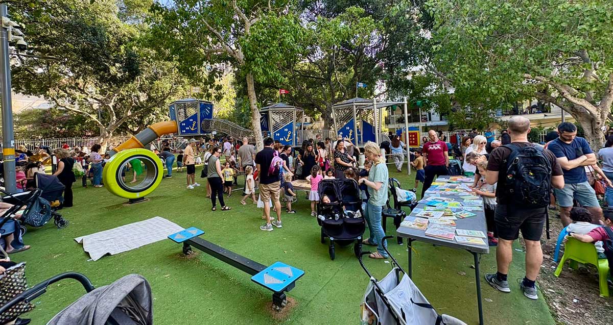 שבוע הקהילה בגבעתיים. קהל רב בגינה ציבורית עם מתקני שעשועים ושולחן ארוך עם ספרים.