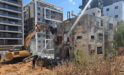 הריסת הבניין האחרון ברמת גן שנפגע מטיל ב"עם כלביא". כלי הנדסי הורס בניין של 4 קומות. לידו אדם מתיז מים על הבניין מצינור וזרנוק.