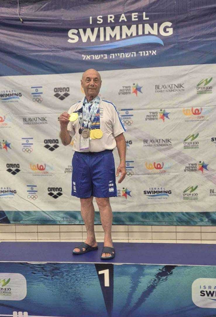 גבר מבוגר עם הרבה מדליות על צווארו מחזיק מדליית זהב ועומד במקום הראשון ומאחוריו שלט של שחייה