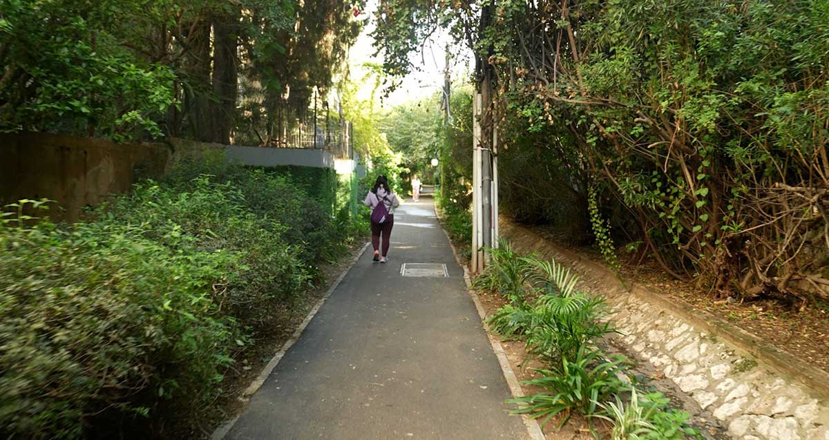 עצים סוככים על שביל הליכה בין בתים בגבעתיים. ילדה פוסעת על השביל בגבה למצלמה