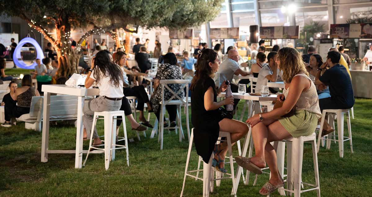 "בציר בעיר" פסטיבל היין 2025. קהל רב יושב ליד שולחנות עם כוסות יין, וגם יש שעומדים עם כוסות יין ביד.